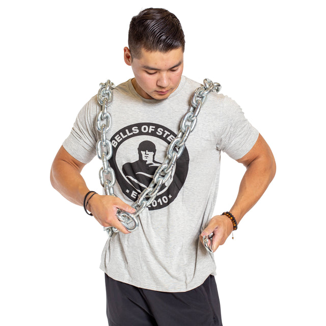 A man with short dark hair wears a gray Bells of Steel t-shirt and black shorts, standing against a white background with the Chain Weight Vest draped over his shoulders as he prepares for intense strength training.