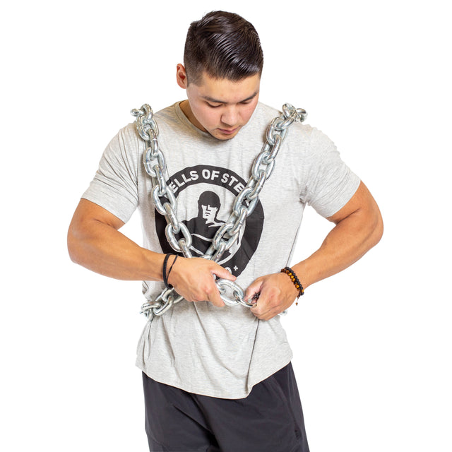 A man wears the Bells of Steel Chain Weight Vest, securing heavy chains around his shoulders and torso as he looks down, standing against a plain white background and preparing for intense strength training.