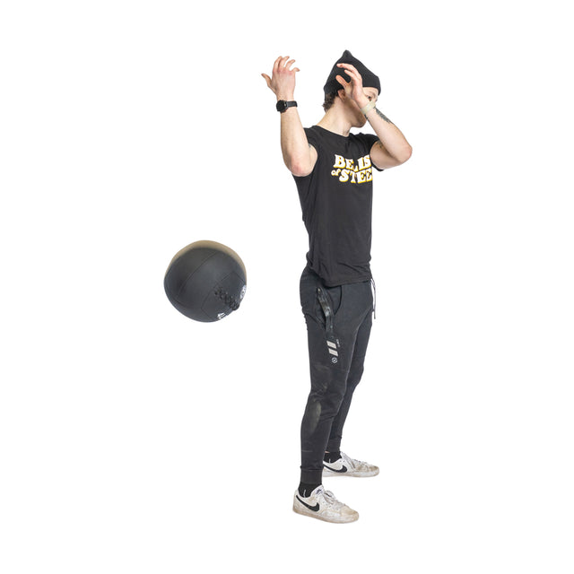 A person in a black T-shirt and pants demonstrates their fitness by tossing a black Triple Stitched Medicine Ball from Bells of Steel. They're wearing a black beanie and white sneakers, all against a white backdrop.