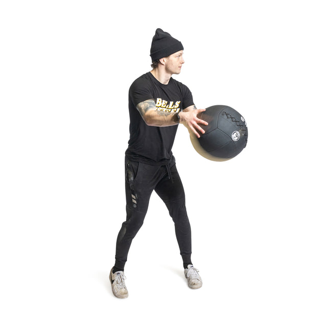 A person wearing a black T-shirt, pants, and beanie expertly balances on one leg while exercising with a Bells of Steel Triple Stitched Medicine Ball against a plain white backdrop, demonstrating varying fitness levels.