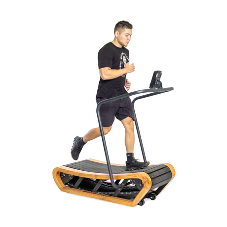 A person is jogging on a Bells of Steel Wooden Residential Manual Treadmill with a curved running surface. Wearing a black T-shirt, shorts, and sneakers, they navigate its compact design effortlessly. The treadmill includes a display screen on the handlebars against a plain white background.