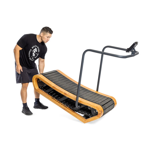 A person in a black t-shirt and shorts adjusts the Bells of Steel Wooden Residential Manual Treadmill, featuring a sleek black surface, wooden frame, and minimalist design with a handlebar device holder.