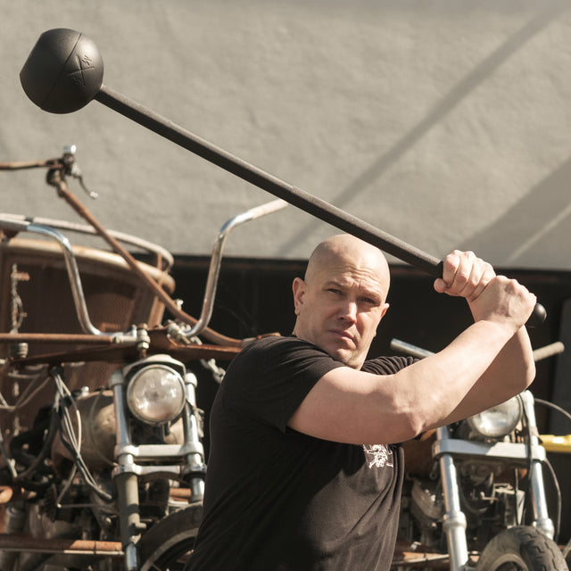In front of parked motorcycles, a person in a black shirt, demonstrating remarkable core strength, wields a Bells of Steel Adjustable Macebell over their shoulder against a beige wall with bold diagonal shadows.