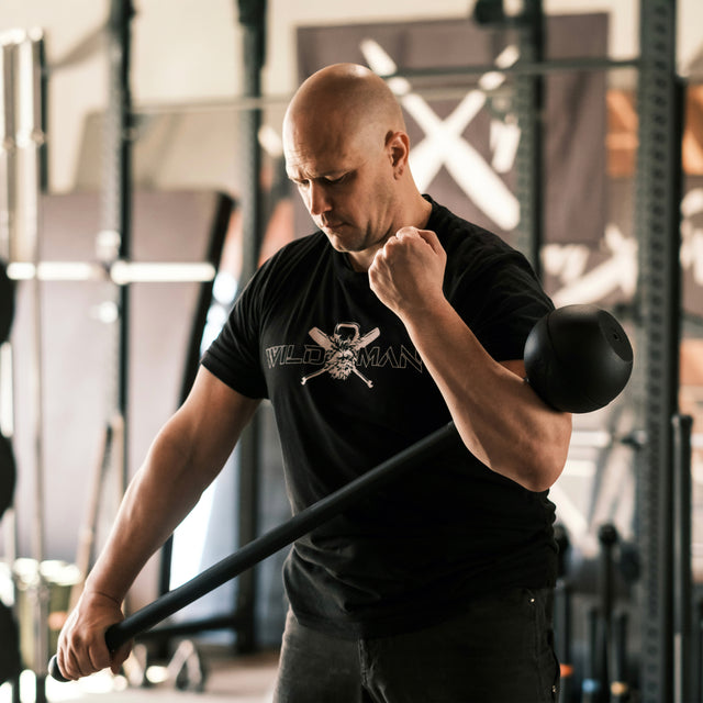 A man in a black "Wildman" shirt skillfully grips a Bells of Steel Adjustable Macebell in the gym. He is focused on enhancing core strength, surrounded by exercise equipment and obscured banners in the background.