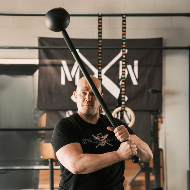 An individual in a black t-shirt performs a core-strength exercise with a Bells of Steel Adjustable Macebell. Amid gym equipment and banners, they maintain focus, enhancing shoulder mobility with each movement.
