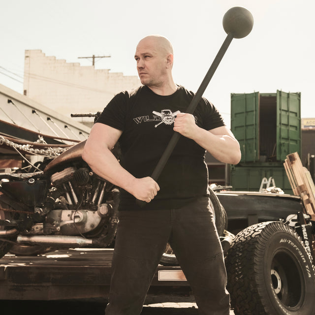 A muscular man in a black T-shirt holds his Bells of Steel Adjustable Macebell beside a motorcycle on a flatbed trailer. With a serious and focused demeanor, he's set to enhance his shoulder mobility and core strength, standing against an industrial area backdrop under broad daylight.