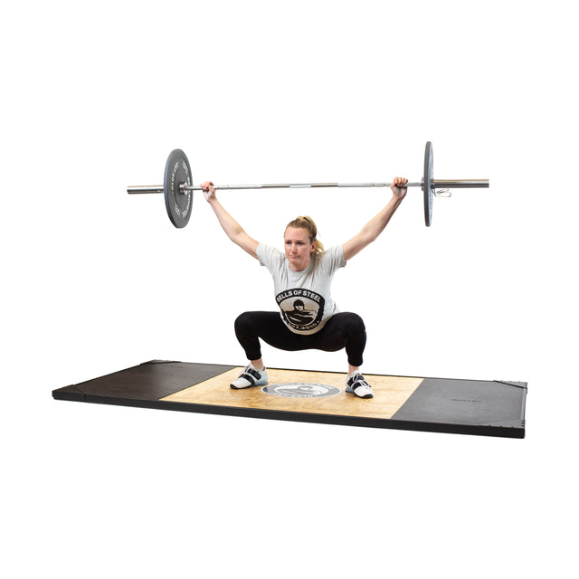 A person performs an overhead squat on a weightlifting platform, using the Bells of Steel Oly Lifting Needle Bearing Bar - Women's, arms fully extended overhead, wearing a gray T-shirt, black pants, and lifting shoes.