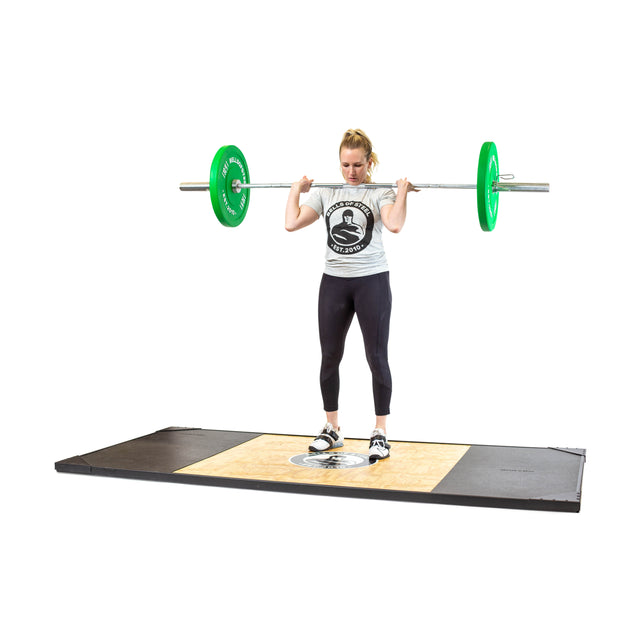 A woman stands on a lifting platform, gripping the Bells of Steel Oly Lifting Needle Bearing Bar - Women's with green weights across her shoulders. She wears black leggings, a gray t-shirt, and weightlifting shoes, preparing for a powerful lift.