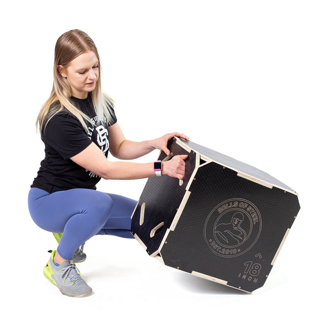 A woman in athletic wear squats while holding and tilting a large black 3 in 1 Anti-Slip Wood Plyo Box by Bells of Steel, featuring an anti-slip surface and “18 inch” marking—ideal for any home gym.