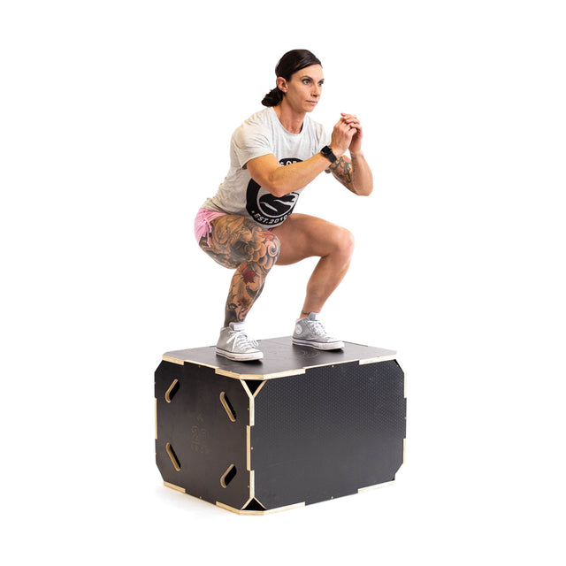 A tattooed woman squats on a Bells of Steel 3 in 1 Anti-Slip Wood Plyo Box, wearing a light t-shirt, pink shorts, and sneakers, with her arms bent and hands clasped in front.