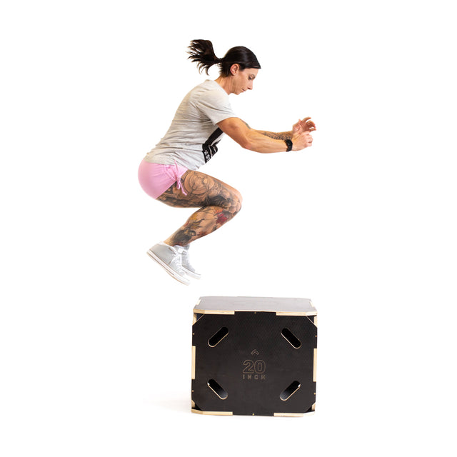 A tattooed person in a gray shirt and pink shorts performs a box jump onto the Bells of Steel 3 in 1 Anti-Slip Wood Plyo Box (20")—ideal for any home gym—against a white background.