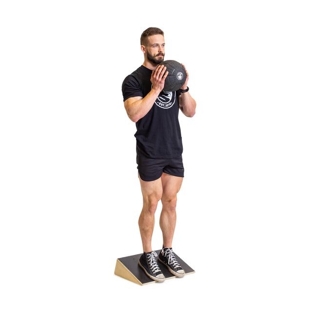 A man in a black outfit stands on a Bells of Steel Squat Wedge Board, holding a weight plate at chest level with both hands as he prepares for an exercise to enhance ankle mobility and squat depth.