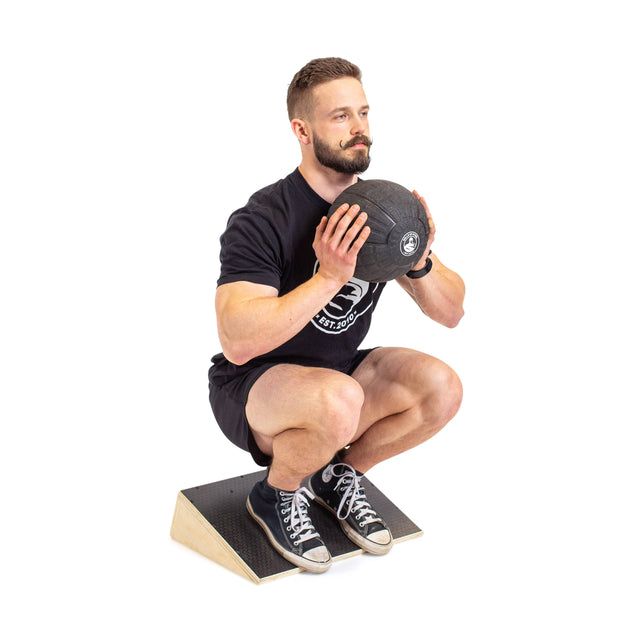 A man in athletic wear uses the Bells of Steel Squat Wedge Board, heels elevated and toes on the floor, holding a weighted ball to his chest against a white background to boost ankle mobility.