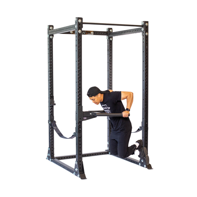 Wearing black attire, an individual exercises with a power rack using Bells of Steel's Y Dip Bar Rack Attachment. Kneeling inside the frame and gripping the horizontal bar, they utilize the adjustable safety arms for versatile workouts.