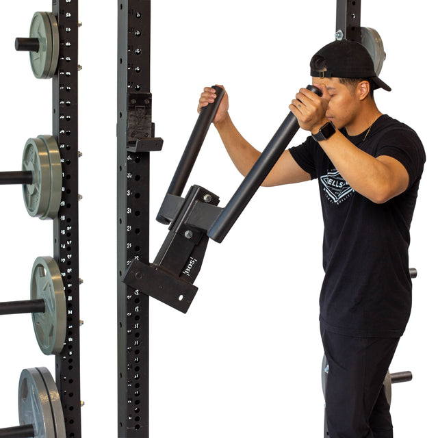 Wearing a black shirt and cap, someone confidently uses the Bells of Steel Y Dip Bar Rack Attachment on a squat rack with a 500lb capacity. Surrounded by weight plates, they grip the handles while standing in profile.