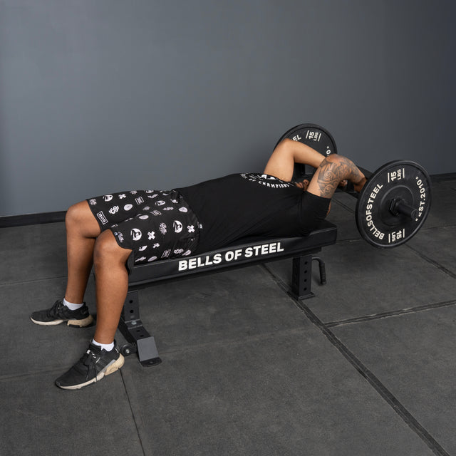 A person lies on a bench labeled “Bells of Steel,” lifting a barbell with bent arms, while an Arch Nemesis Tricep Bar from Bells of Steel rests nearby in the gym with gray walls and black flooring.