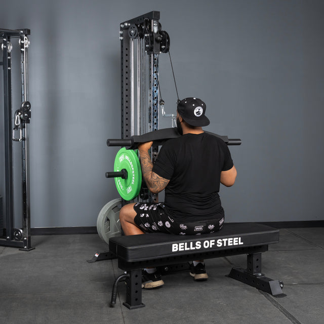 A person in black attire uses the Arch Nemesis Tricep Bar by Bells of Steel to perform a lat pulldown while seated on a branded gym bench.