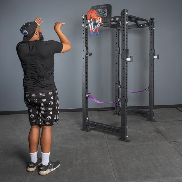 A man in a black shirt, patterned shorts, and a cap shoots a basketball toward the Bells of Steel Rack Attached Basketball Hoop w/ Ball in a gym.