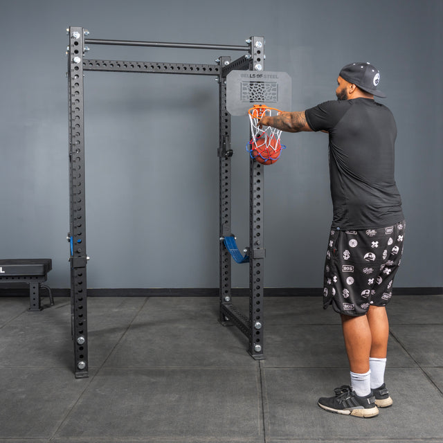 In a home gym with gray walls and black flooring, a man in athletic wear loads a barbell onto a squat rack, while the Bells of Steel Rack Attached Basketball Hoop w/ Ball adds versatility to his setup.