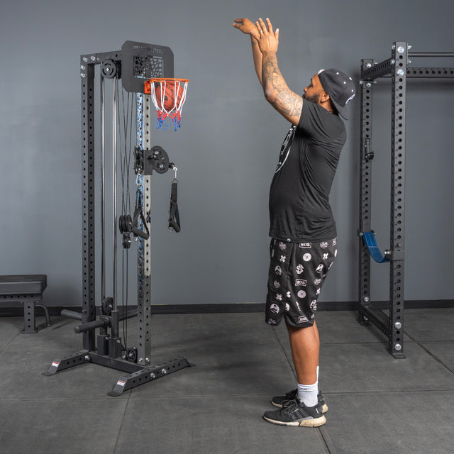 A man in athletic wear pretends to shoot a ball into the Bells of Steel Rack Attached Basketball Hoop w/ Ball, mounted on a gym cable machine, with exercise equipment visible in the background.