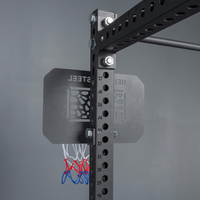 A close-up of the Bells of Steel Rack Attached Basketball Hoop w/ Ball, featuring a black metal adjustable frame, compact steel backboard with "BELLS OF STEEL" cut out, and a red, white, and blue net against a gray background.