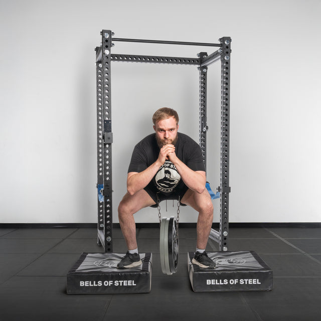 A man performs a belt squat with the Bells of Steel USA Dip Belt, standing on two raised Bells of Steel platforms in a gym with a black mat floor and white walls.