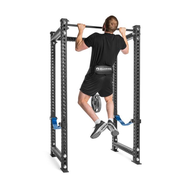 A person wearing athletic clothing and a Bells of Steel USA Dip Belt does a pull-up on a black power rack with safety straps, highlighting bodyweight training, isolated on a white background.