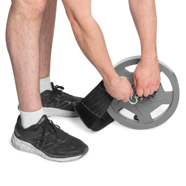 A person wearing black sneakers attaches the Bells of Steel USA Dip Belt, featuring reinforced stitching, to a gray weight plate against a white background, preparing for bodyweight training.