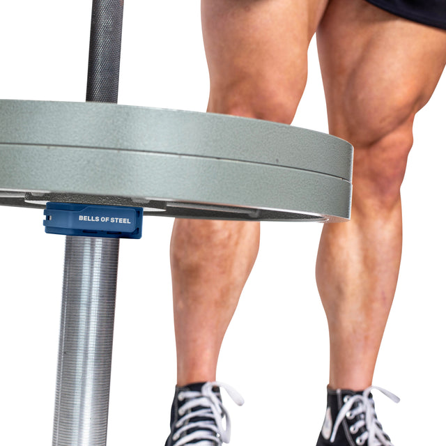 Close-up of a personโs muscular legs and feet in black sneakers standing behind a barbell loaded with weight plates, secured using Bells of Steel Magnetic Clamp Collars.