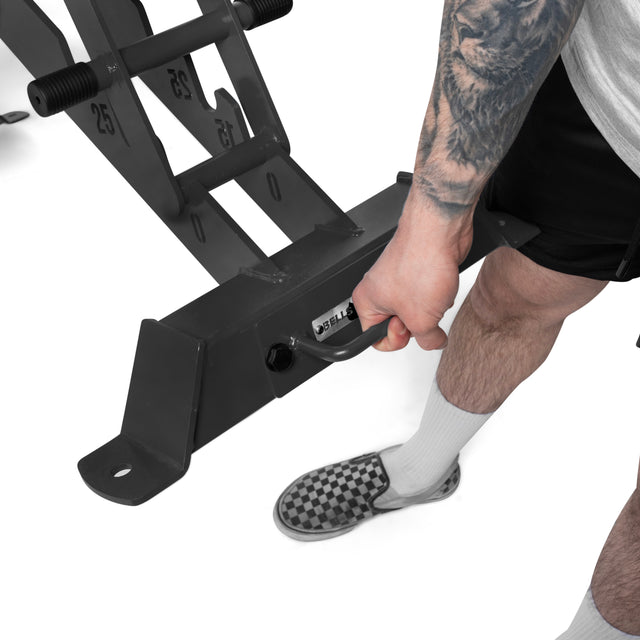 A person with a detailed forearm tattoo grips the handle of the Bells of Steel Buzz-Saw Heavy-Duty Adjustable Bench while wearing checkered slip-ons, white socks, black shorts, and a gray shirt.