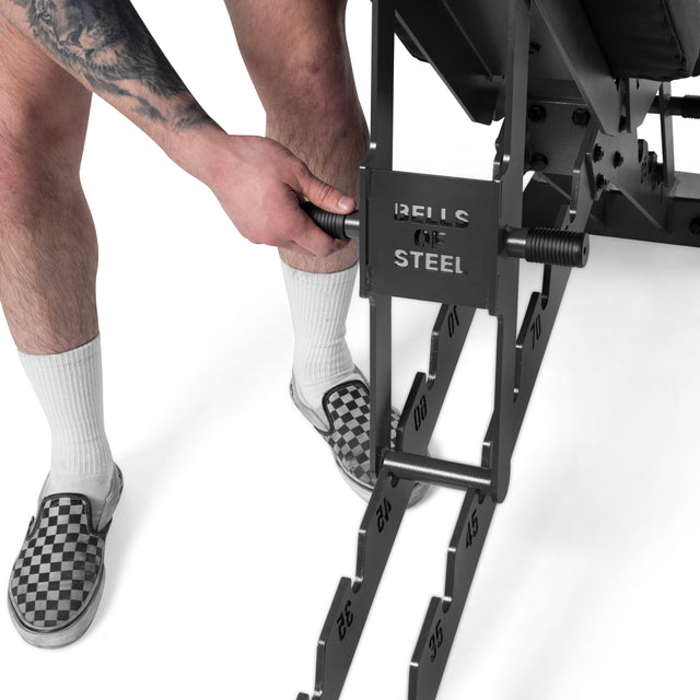 A person adjusts the incline on a Bells of Steel Buzz-Saw Heavy-Duty Adjustable Bench. Only their legs in white socks, black-and-white checkerboard slip-on shoes, and a tattooed arm are visible.
