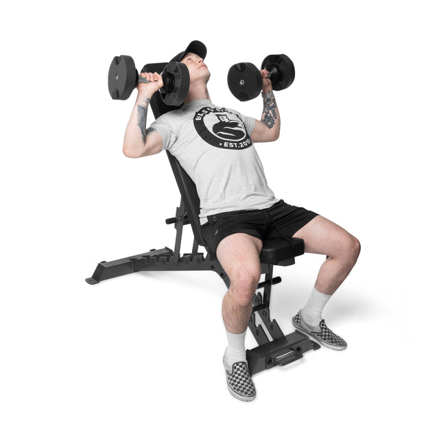 Wearing a gray t-shirt, black shorts, and checkered shoes, a person performs an incline dumbbell press on a Bells of Steel Buzz-Saw Heavy-Duty Adjustable Bench against a plain white background.