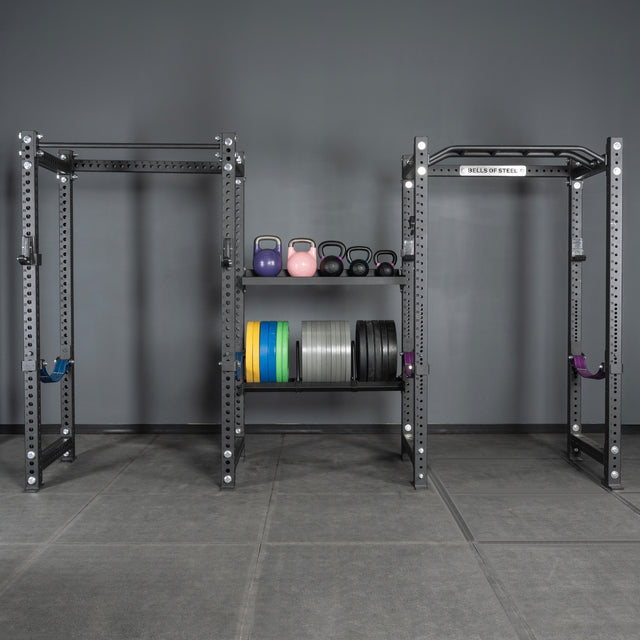 A gym setup featuring two squat racks, a pull-up bar, colorful kettlebells, and a Bells of Steel USA rack with plates—neatly organized with Divider for Bumper Plate Shelf (Pack of 3) against a gray wall and floor with shelving.