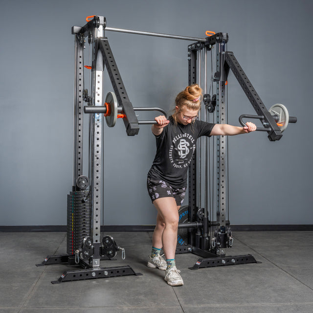 Wearing athletic gear, a person uses the Bells of Steel Lever Arms Rack Attachment—loaded with Olympic plates—on a squat rack. The gym has gray walls/flooring, an adjustable pull-up bar, and other fitness equipment in view.
