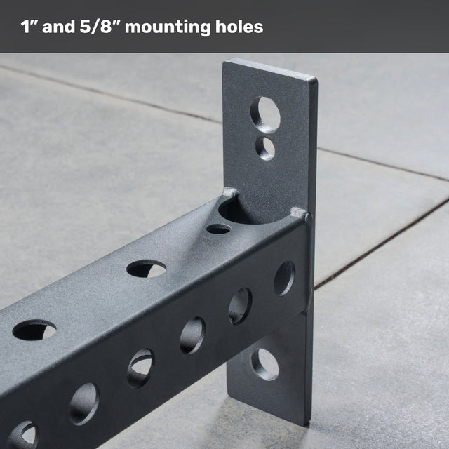 A close-up of the Bells of Steel USA Crossmember For Converter Bench - 43", a black metal bracket with multiple 1” and 5/8” circular mounting holes, shown attached at a right angle on a concrete floor for versatile rack setups.
