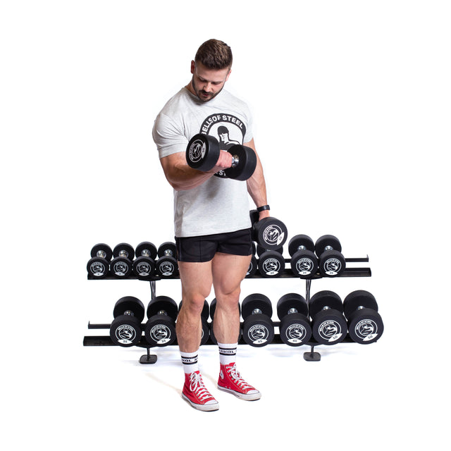 A man in a white T-shirt, black shorts, and red sneakers lifts a dumbbell in a gym, standing before the Bells of Steel Commercial Interchangeable Weight Rack filled with various weights.