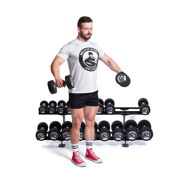 A man in a gray shirt and black shorts lifts dumbbells beside a Bells of Steel Commercial Interchangeable Weight Rack in the gym. He wears red sneakers and white socks.