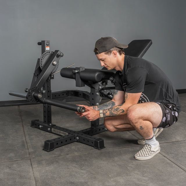 A man in a black tee, patterned shorts, and backward cap crouches by a Bells of Steel USA Legacy Leg Extension / Hamstring Curl Machine - Plate Loaded, adjusting its settings in a fitness studio with gray walls and flooring.