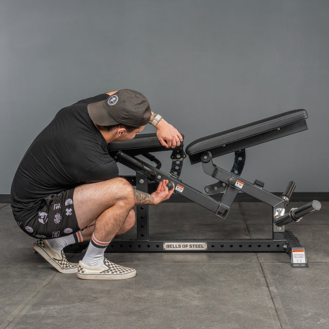 A person in casual clothes and a cap adjusts the angle of a black Legacy Leg Extension / Hamstring Curl Machine - Plate Loaded by Bells of Steel USA in a home gym with a gray background.