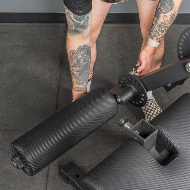 A person with leg tattoos adjusts the settings on a Legacy Leg Extension / Hamstring Curl Machine - Plate Loaded by Bells of Steel USA, focusing on the adjustment dial and padded rollers—perfect for any home gym.