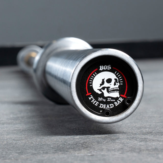 Close-up of a Bells of Steel USA Deadlift Bar end cap with a skull graphic and "BOS The Dead Bar 20 kg 27 mm" text, resting on a gray floor—ideal for serious powerlifting or strongman training.