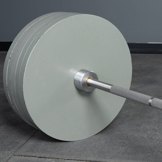 A Bells of Steel USA Deadlift Bar with multiple large gray weight plates sits on a gym floor, set against a gray wall and black baseboard, capturing the serious atmosphere of powerlifting and strongman workouts.
