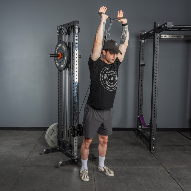 Wearing a black t-shirt, shorts, and cap, a person stands in the gym using the Bells of Steel USA Flat-Iron Tricep Press Cable Attachment for overhead triceps extensions, arms extended to maximize tricep activation.