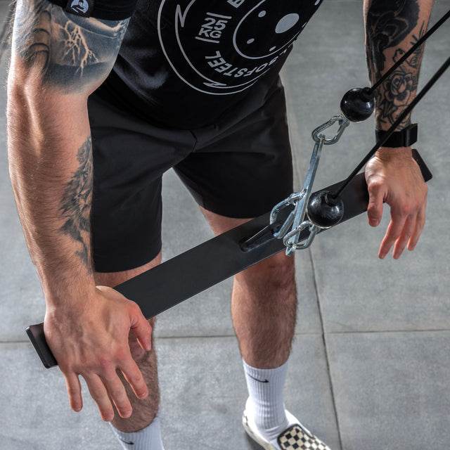 A tattooed individual uses the Bells of Steel USA Flat-Iron Tricep Press Cable Attachment, gripping the straight bar while wearing a black shirt, black shorts, white socks, and checkered sneakers on a gray gym floor.