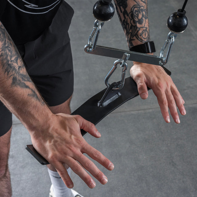 A person with tattooed arms grips the Bells of Steel USA Flat-Iron Tricep Press Cable Attachment in a gym, ready to work out. They're dressed in black shorts, white socks, and athletic shoes.
