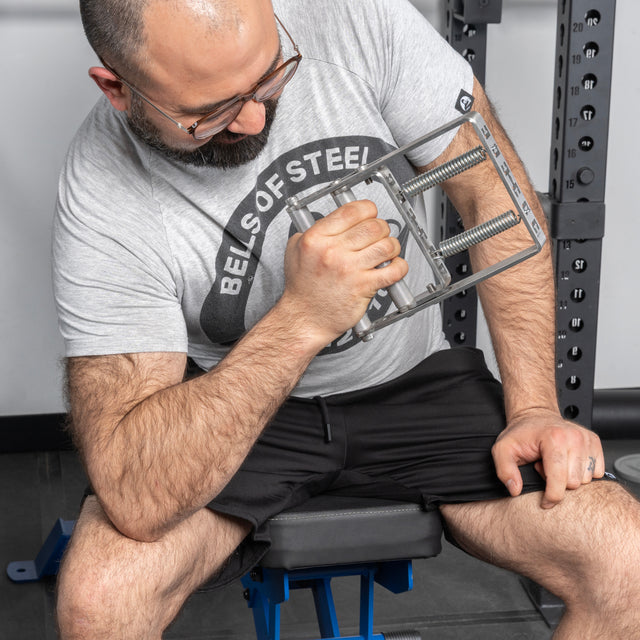 A bearded man in glasses and a “Bells of Steel USA” shirt sits on a bench, intently using the Adjustable Forearm Heavy Trainer with his right hand to boost grip strength and improve his exercise routine.