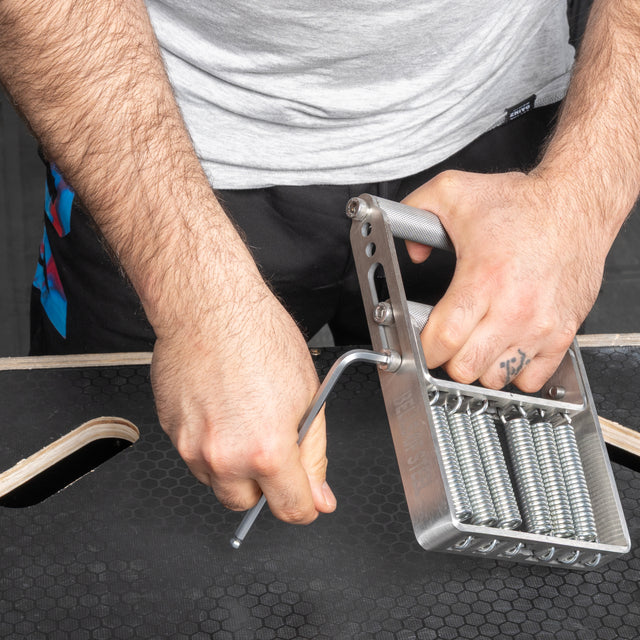 A person uses a hex key to adjust the tension on the Bells of Steel USA Adjustable Forearm Heavy Trainer, which has multiple visible springs showing its adjustable resistance, placed on a black surface.