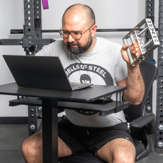 A man in a "Bells of Steel" t-shirt and black shorts stands at a gym desk, using a laptop while holding the Bells of Steel USA Adjustable Forearm Heavy Trainer, surrounded by gym equipment featuring adjustable resistance.