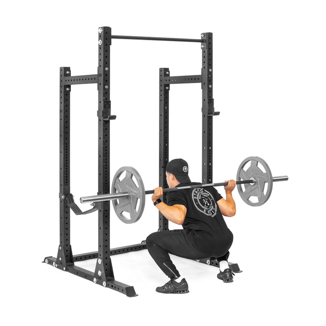 A person in black athletic wear and a cap is performing a barbell squat in their home gym, using the Bells of Steel Hydra Half Rack - Prebuilt (3" x 3", ⅝" Holes), with a weighted barbell across their shoulders.