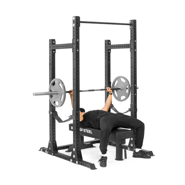 A person in black athletic wear prepares to bench press on a Bells of Steel Hydra Half Rack - Prebuilt (3" x 3", ⅝" holes), gripping a barbell while lying on a bench in their home gym.